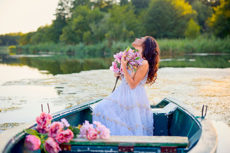 Beautiful woman in white dress on a boat with flowers on a lake in the evening light.の写真素材