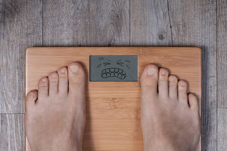 Heavy weight - display shows emoticon face suffering reaction. Legs of man standing on weight scale.の写真素材