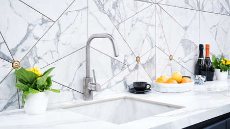 The interior of the modern kitchen is illuminated with a gray stone countertop with a luxury washbasin and mixer, fruit orange and flowers, a bottle with red wine, champagne and chocolateの写真素材