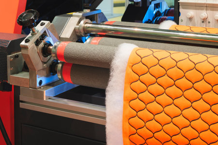 A machine is making a piece of orange fabric with a pattern of squares. The machine is made of metal and has a black handleの写真素材