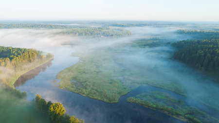 A foggy morning over a river with trees in the background. The sky is a light blue colorの写真素材