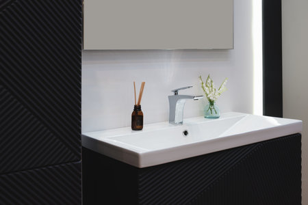 A bathroom sink with a vase of flowers and a bottle of essential oil on the counter. The sink is white and the counter is blackの写真素材