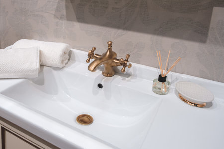 A sink with a gold faucet and a white towel on top of it. A vase with a few sticks in it sits on the counter next to the sinkの写真素材