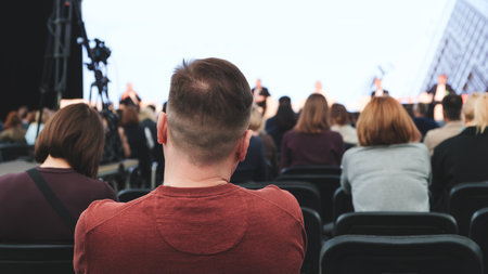 A man in a red shirt sits in a row of chairs in a large auditorium. The audience is attentive and engaged, listening to a speaker. The atmosphere is serious and focusedの写真素材