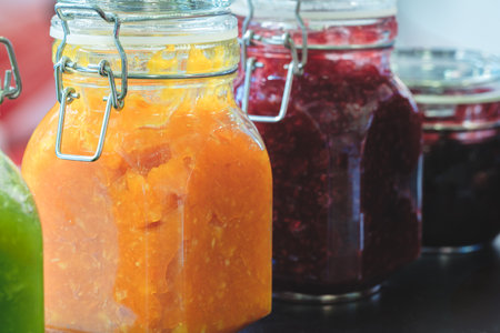 A row of jars with different colored jams, including orange, purple, and green. The jars are lined up on a tableの写真素材