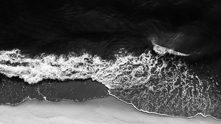aerial view from drone, black and white photograph of wave crashing onto beach. wave is large and...