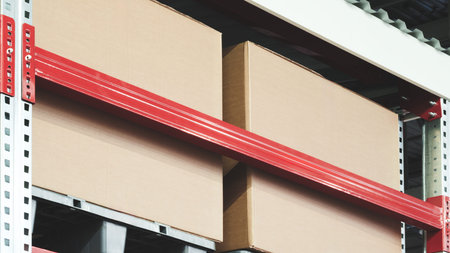 Row of cardboard boxes are stacked on a shelf. The boxes are brown and red. The shelf is made of...
