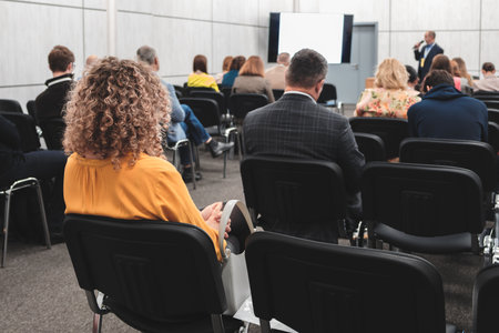 A businesswoman sits in a conference room full of people. A male speaker in a suit stands in front of a projection screen with a microphone. Business education presentation training concept.の写真素材