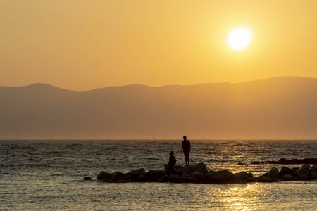 Silhouettes Of Men Fishing On Rocks At Sunsetの写真素材