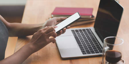 Close-up of female hand holding blank screen smartphone in modern comfortable workspace with laptop computer and office suppliesの写真素材