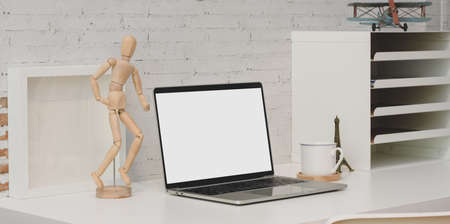 Contemporary workplace with blank screen laptop, wooden figure on white table and brick wall backgroundの写真素材