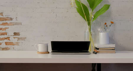 Minimal comfortable workplace with laptop and office supplies on white wooden desk and brick wall backgroundの写真素材
