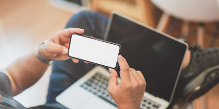 Young male freelancer working on his project with laptop computer while using blank screen smartphoneの写真素材