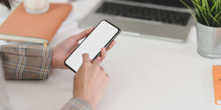 Close-up view of young businesswoman holding blank screen smartphone while working on laptop computerの写真素材