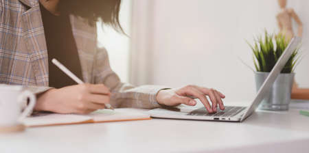 Motivated young businesswoman planing her strategy while typing on laptop and writing on notebook