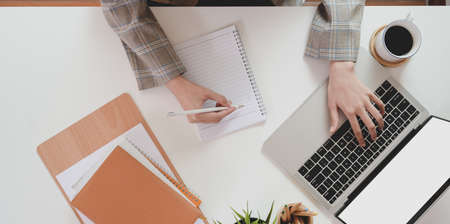Top view of businesswoman typing on laptop computer and writing her idea on notebook in comfortable...