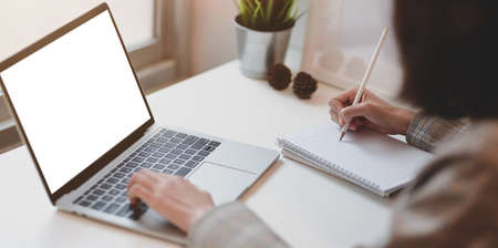 Young businesswoman typing on blank screen laptop computer and writing her idea on notebook