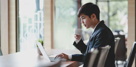Young professional businessman drinking a cup of coffee while working with laptop in office roomの写真素材