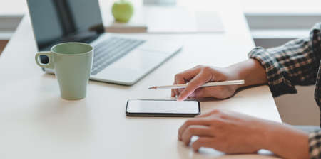 Young male freelancer looking at blank screen smartphone while working on laptop computer