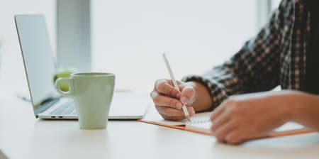 Close-up of male freelancer writing his ideas on notebook in minimal home-officeの写真素材