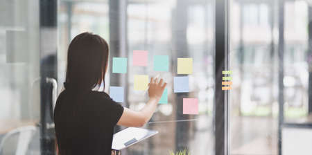 Young female graphic designer collecting ideas from sticky notes on the glass wall in office room
