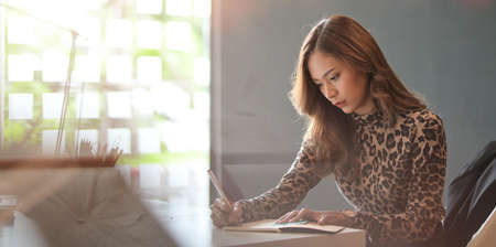 Attractive young businesswoman working on her project and writing her idea in trendy office roomの写真素材