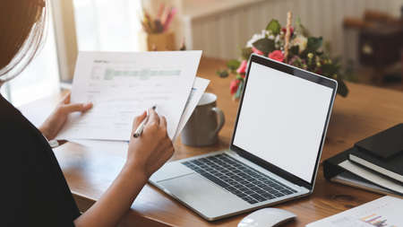 Close-up view of businesswoman working on her project with blank screen laptop computer while looking at the paperの写真素材