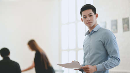 Attractive young professional businessman looking at the camera while holding document and standing in the meeting roomの写真素材