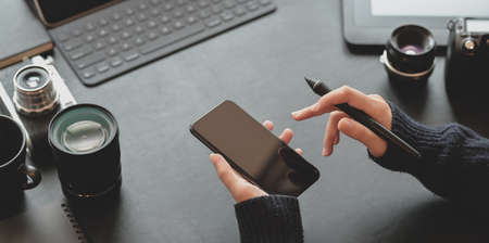 Close-up view of female photographer holding smartphone in dark stylish workplace with camera and office suppliesの写真素材