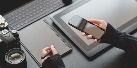 Cropped view of female photographer holding smartphone in dark stylish workplace with office supplies on black leather deskの写真素材