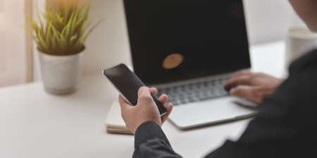 Close-up of young male holding smartphone while typing on laptop computer in comfortable workplace