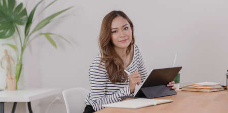 Portrait of beautiful designer working on her project with tablet and smiling at the camera in modern workplace with office suppliesの写真素材