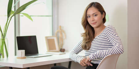 Beautiful female designer editing her project with laptop and looking at the camera in comfortable workplace near the windowの写真素材