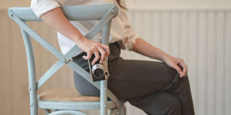Cropped view of young female photographer holding vintage camera while sitting on the chair in comfortable roomの写真素材