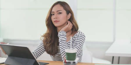 Portrait of beautiful designer working on her project with tablet and looking at the camera in modern workplace with office suppliesの写真素材
