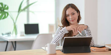 Beautiful female designer editing her project with tablet and smiling at the camera in comfortable workplace with decorationsの写真素材