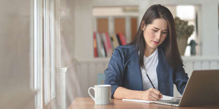 Professional female freelancer working on her project while writing on notebook in modern workplaceの写真素材