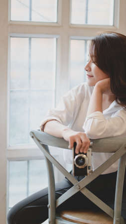 Young beautiful photographer holding camera while sitting on the chair in her modern workplace