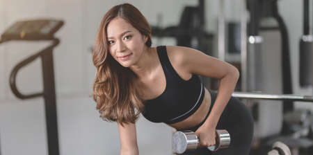 Fit asian athletic woman lifting weights inside home gym and smiling to the camera, building muscle conceptsの写真素材