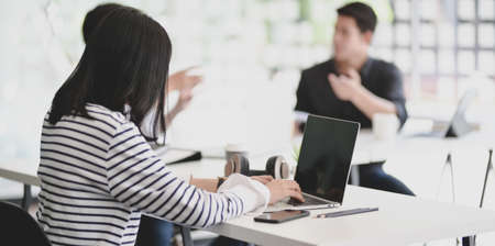 Young female freelancer typing on laptop computer about the meeting details with her male coworkers in the backgroundの写真素材