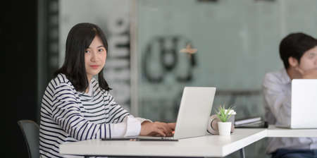Young female freelancer working on her project with laptop computer and smiling to camera, with her male coworker in the backgroundの写真素材
