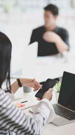 Young female freelancer looking for information on smartphone with her male coworker in the backgroundの写真素材
