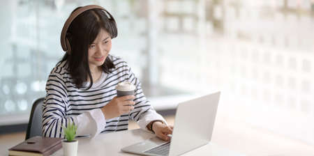 Cheerful young female freelancer typing on laptop computer while drinking a cup of coffee in her workplaceの写真素材