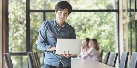 Young male professional UX web developer standing in the meeting room and holding laptop computerの写真素材