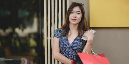 Portrait of beautiful asian woman holding her smartphone and shopping bag after shopping at the mallの写真素材
