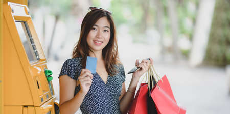 Young asian woman holding credit card and shopping bag after withdrawing the cash from ATM machineの写真素材
