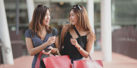 Two beautiful asian women chatting together and enjoying their free time at the shopping mall while holding shopping bagの写真素材