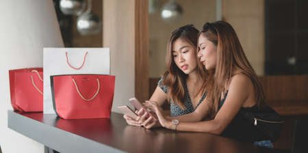 Two beautiful asian women checking their smartphone and chatting together after shopping in the mallの写真素材