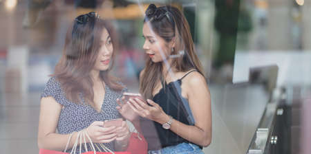 Two beautiful asian women chatting together and holding smartphone while shopping at the mallの写真素材