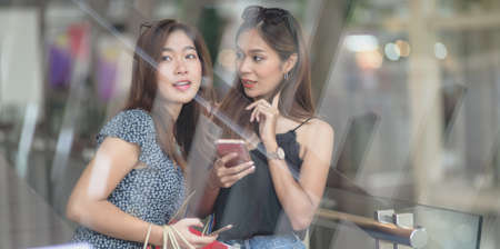 Two beautiful asian women chatting together and holding smartphone while shopping at the mallの写真素材